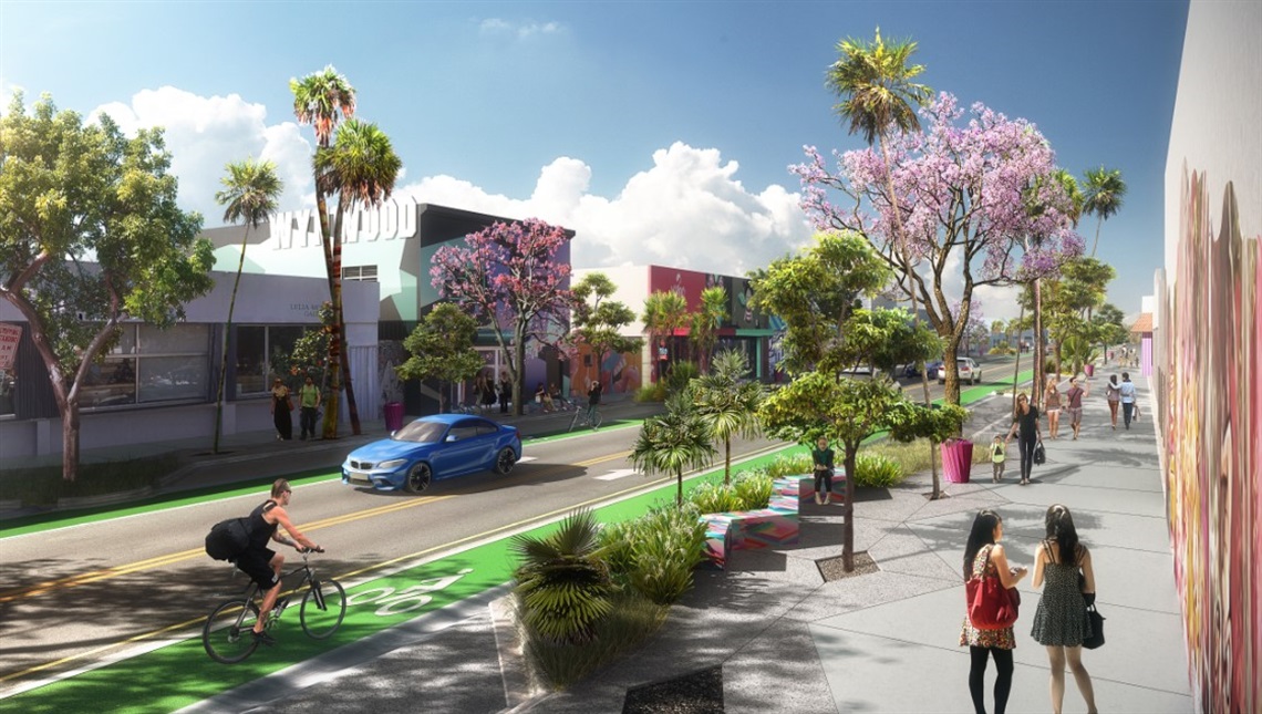Street view showing buildings on the left with trees on the sidewalk, cars driving along the street, a person riding a bike on the bike path, and on the right part, a sidewalk with trees and people walking, near a colorful mural